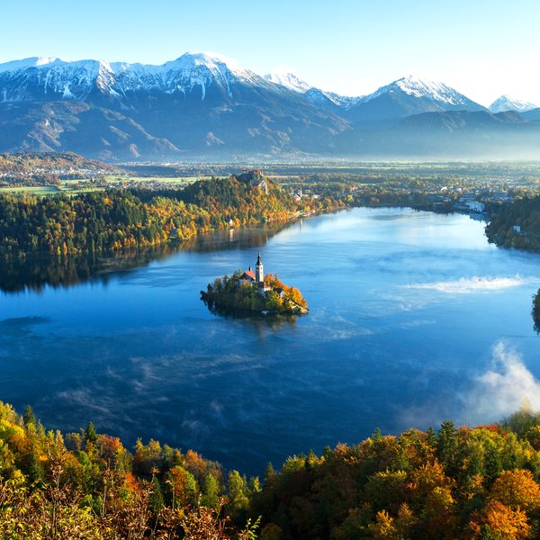 Ein malerischer Blick auf den Bleder See, umgeben von buntem Herbstlaub und majestätischen Bergen im Hintergrund.
