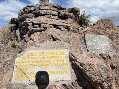 Ein Kind berührt eine Tafel mit der Aufschrift zur Cruz del Condor und blickt auf die monumentale Felsformation.