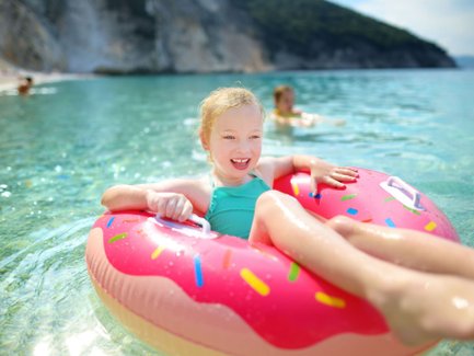 Ein fröhliches Mädchen mit blonden Locken genießt das Wasser in einem bunten Donut-Floatie an einem sonnigen Strand.