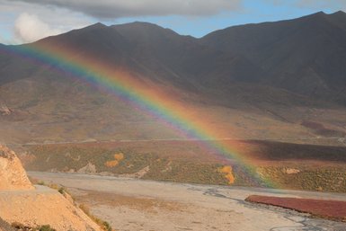 Ein strahlender Regenbogen spannt sich über eine karge, farbenfrohe Landschaft mit Bergen im Hintergrund.