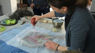 Eine Frau arbeitet an einem Tisch, während ein Mann und ein Kind zuschauen, während sie mit Textilien experimentiert.