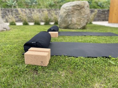 Auf einer grünen Wiese liegen zwei schwarze Yogamatten und zwei Korkblöcke, umgeben von einer ruhigen, natürlichen Umgebung.