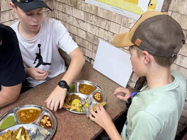 Zwei Jungen sitzen an einem Tisch und genießen indisches Streetfood von einem runden Metalltablett mit verschiedenen Gerichten.