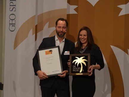 Zwei Personen stehen auf einer Bühne und halten Auszeichnungen, während im Hintergrund ein großes Palmbaum-Logo zu sehen ist.