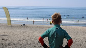 Ein Junge in einem grünen Surfanzug beobachtet das Meer, während andere im Wasser surfen und spielen.