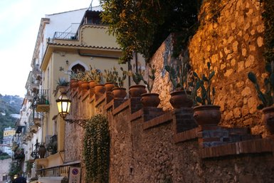 Eine malerische Gasse in Taormina, gesäumt von Töpfen mit Kakteen und warmem Licht, das die Steine beleuchtet.