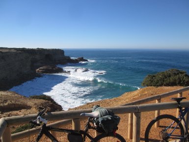 Zwei Fahrräder stehen an einem Geländer mit Blick auf die Wellen, die gegen die Klippen schlagen, unter klarem Himmel.