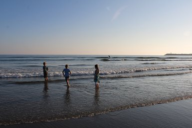 Drei Kinder stehen im flachen Wasser, während sanfte Wellen an den Strand rollen und die Sonne am Horizont scheint.
