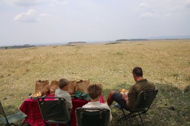 Drei Personen sitzen auf Campingstühlen und genießen ein Picknick mit Blick auf eine weite, grüne Landschaft.