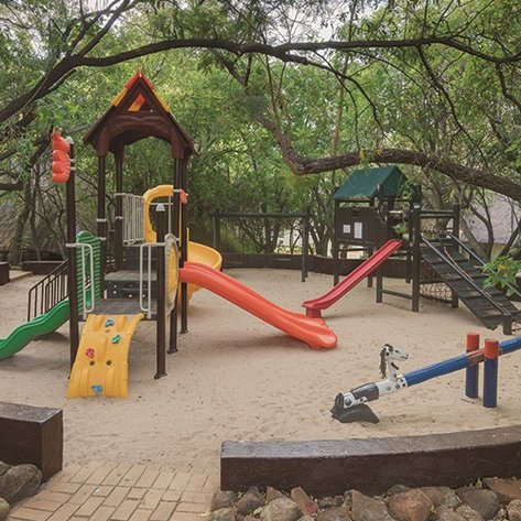 Ein Spielplatz im Freien, umgeben von Bäumen, mit Rutschen, Klettergerüsten und einem Sandbereich für Kinder.