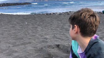 Ein Junge sitzt am Strand und gräbt mit seinen Händen im schwarzen Sand, während er auf das Meer blickt.