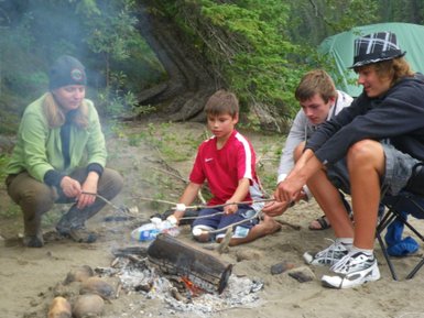 Eine Gruppe von vier Jugendlichen sitzt um ein Lagerfeuer und röstet Marshmallows auf langen Stöcken.