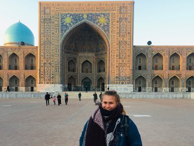 Eine Frau steht lächelnd vor der beeindruckenden Architektur des Registan in Samarkand, umgeben von Besuchern.