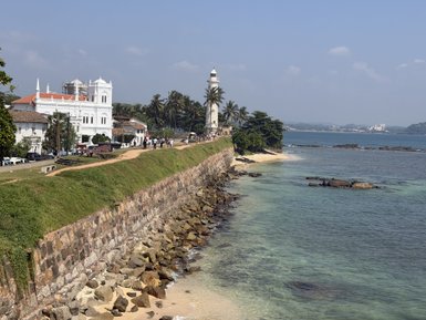 Ein malerischer Küstenabschnitt mit einem Leuchtturm und historischen Gebäuden, umgeben von Palmen und klarem Wasser.