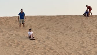 Ein Mädchen sitzt auf einem Schlitten und rutscht eine Sanddüne hinunter, während ein Mann hinter ihr steht.
