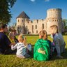 Eine Familie sitzt auf dem Gras vor einer alten Burg, während sie sich unterhält und die Sonne genießt.