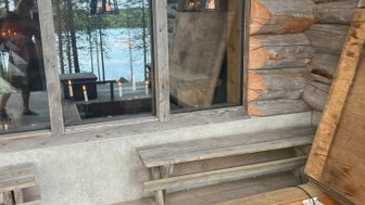 Ein rustikales Holzhaus mit großen Fenstern bietet einen Blick auf einen ruhigen See, umgeben von hohen Bäumen.