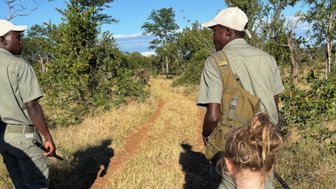 Zwei Männer in khakifarbener Kleidung gehen auf einem schmalen Pfad durch die Natur, während ein Kind mit einem Rucksack folgt.