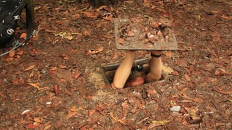 Ein Mensch hebt eine schwere Klappe von einem versteckten Tunnel im Boden, umgeben von herbstlichen Blättern.