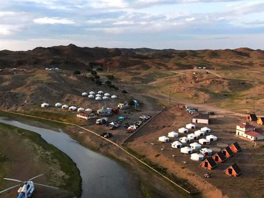 Eine Luftaufnahme zeigt ein Camp in der mongolischen Landschaft mit Jurten und einem Fluss, umgeben von sanften Hügeln.