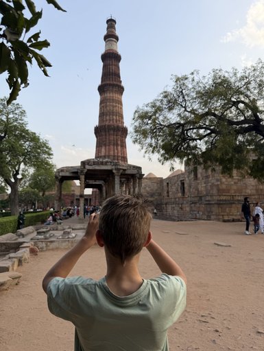 Ein Junge steht mit dem Rücken zur Kamera und fotografiert den beeindruckenden Qutub Minar, umgeben von historischen Ruinen.