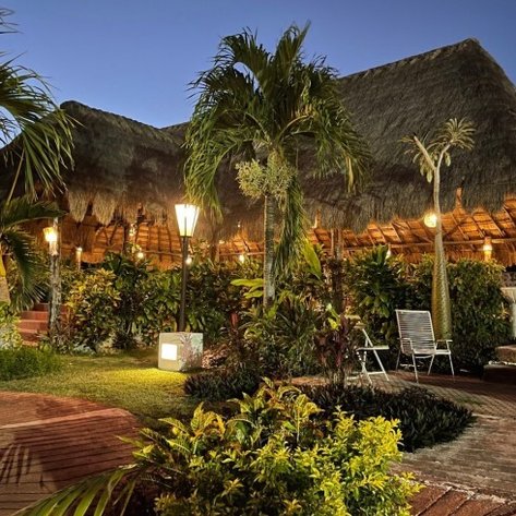 Eine tropische Landschaft mit einem strohgedeckten Gebäude, umgeben von üppigem Grün und sanfter Beleuchtung in der Dämmerung.