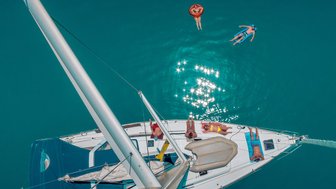 Eine Gruppe von Menschen entspannt auf einem Segelboot, während andere im glitzernden Wasser schwimmen.
