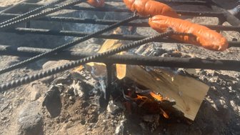 Über dem offenen Feuer brutzeln mehrere Würstchen auf einem Grillrost, während Rauch in die Luft steigt.