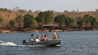 Eine Gruppe von fünf Personen genießt eine Bootsfahrt auf einem ruhigen Gewässer, umgeben von grünen Bäumen und trockenem Gelände.