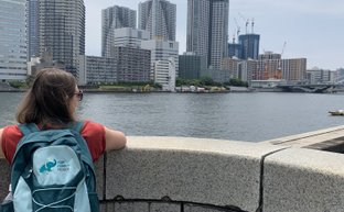Eine Person mit einem Rucksack blickt über das Wasser auf eine moderne Stadtlandschaft mit hohen Gebäuden.