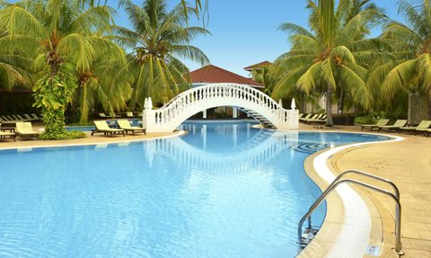 Ein malerischer Pool mit einer eleganten weißen Brücke, umgeben von üppigen Palmen und Sonnenliegen, strahlt tropisches Flair aus.