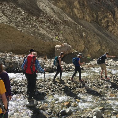 Eine Gruppe von Wanderern überquert einen glitzernden Bach, umgeben von steinigen Ufern und hohen Felsen.