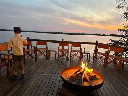 Ein Junge steht am Ufer eines ruhigen Gewässers, während ein Lagerfeuer in der Nähe flackert und der Sonnenuntergang den Himmel färbt.