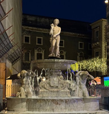 Eine beeindruckende Statue eines jungen Mannes steht in der Mitte eines beleuchteten Brunnens, umgeben von sprudelndem Wasser.