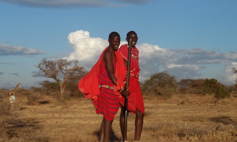 Zwei fröhliche Maasai-Krieger in leuchtend roten Gewändern posieren in der afrikanischen Savanne unter einem blauen Himmel.