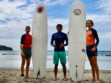 Drei junge Surfer stehen mit ihren Brettern am Strand von Playa Samara, umgeben von sanften Wellen und blauem Himmel.