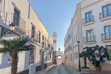 Eine ruhige Straße mit weißen Gebäuden, Palmen und einem markanten Leuchtturm im Hintergrund, der den Blick anzieht.