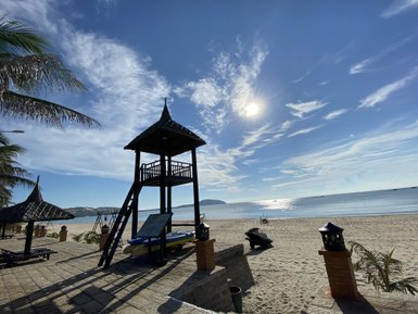 Ein Rettungsturm steht am Strand, umgeben von Palmen und einem klaren blauen Himmel, während die Sonne hell scheint.