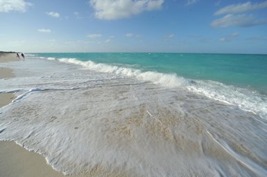 Sanfte Wellen brechen an einem goldenen Sandstrand unter einem strahlend blauen Himmel mit wenigen Wolken.