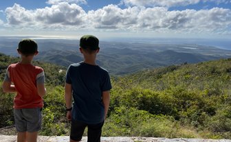 Zwei Jungen stehen auf einem Aussichtspunkt und blicken auf eine weite, bewaldete Landschaft unter einem bewölkten Himmel.