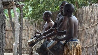 Drei Männer spielen leidenschaftlich auf traditionellen Trommeln, umgeben von einer natürlichen Kulisse und einem Zaun aus Zweigen.