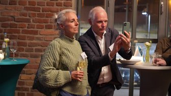Eine Frau mit grauen Haaren in einem grünen Pullover hält ein Glas Sekt, während ein Mann sie mit einem Smartphone fotografiert.