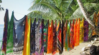 Bunte Tücher hängen an einer Leine, umgeben von Palmen und dem ruhigen Wasser eines Strandes im Hintergrund.