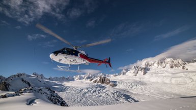 Ein Hubschrauber schwebt über dem Franz-Josef-Gletscher, umgeben von schneebedeckten Bergen und klarem blauen Himmel.