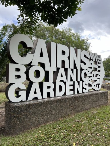 Ein großer, moderner Schriftzug mit den Worten 'Cairns Botanical Gardens' steht im Freien, umgeben von grünen Pflanzen und Bäumen.