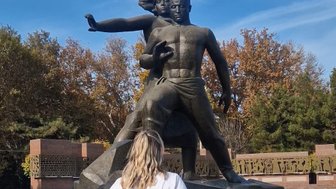 Eine Frau steht vor einem beeindruckenden Denkmal, das einen mutigen Mann und eine Frau darstellt, umgeben von herbstlichen Bäumen.