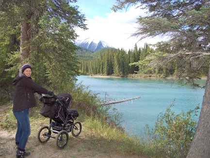 Eine Frau mit einem Kinderwagen steht am Ufer eines klaren Flusses, umgeben von hohen Bäumen und Bergen im Hintergrund.
