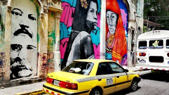 Bunte Wandmalereien zeigen Porträts von Menschen in einem urbanen Umfeld, während ein gelbes Taxi vorbeifährt.
