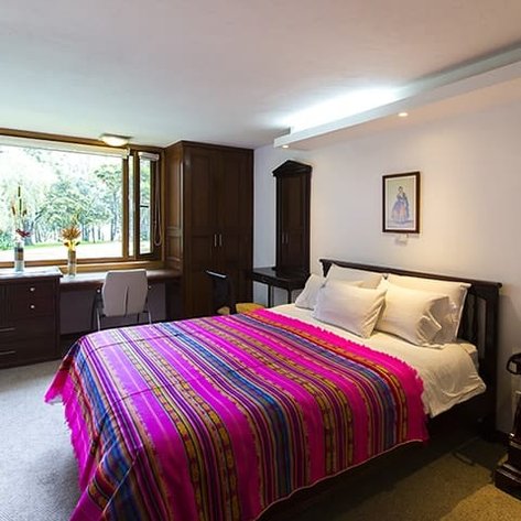 Ein gemütliches Schlafzimmer mit einem bunten Bettbezug und großen Fenstern, die auf einen grünen Garten blicken.