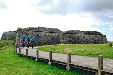 Drei Radfahrer in blauen Trikots fahren auf einem Holzsteg, umgeben von einer alten Steinmauer und grünem Gras.